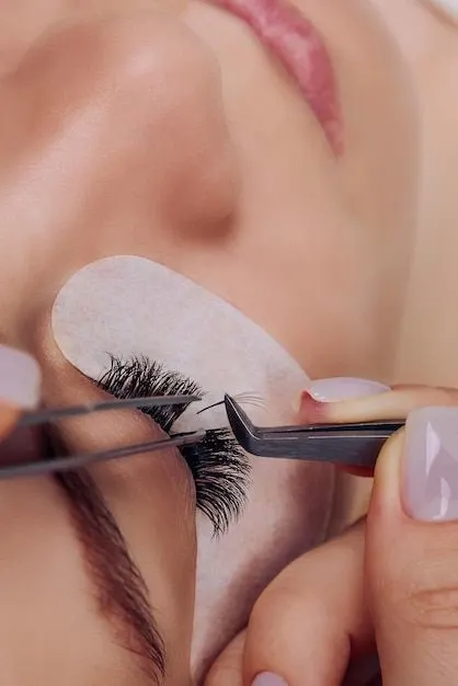 A close-up view of a beauty technician applying individual eyelash extensions to a woman's closed eye using tweezers, with an under-eye pad in place.